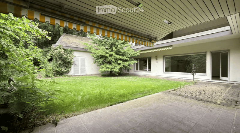 A well-maintained garden area with a paved pathway leading to a modern house. The house features large windows, a covered entrance, and a small shed. The garden is lush with green grass and various plants.