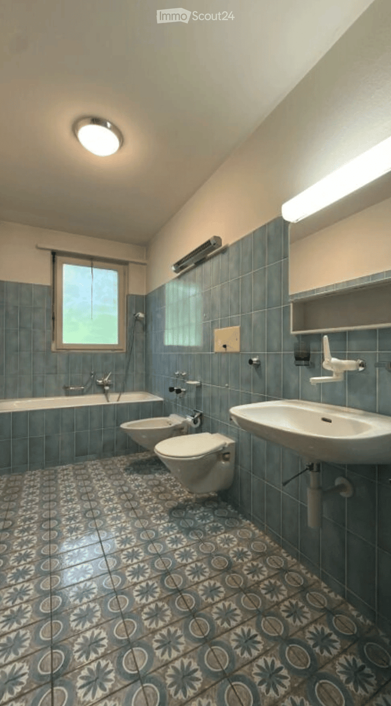 Bathroom with blue tiles, a bathtub, toilet, bidet, sink, and a window. The floor features a patterned tile design.