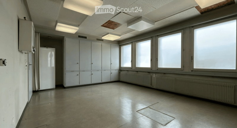 Empty room with white walls, large windows, built-in cabinets, and a refrigerator. The floor is tiled, and the ceiling has fluorescent lighting.