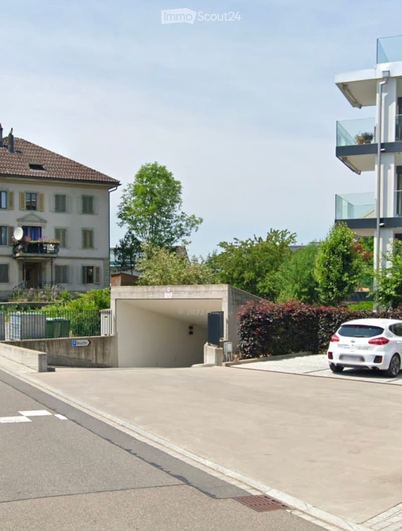 A modern residential complex with a multi-story building, a balcony, and a covered parking area. A white car is parked near the entrance, and there are trees and shrubs surrounding the area.