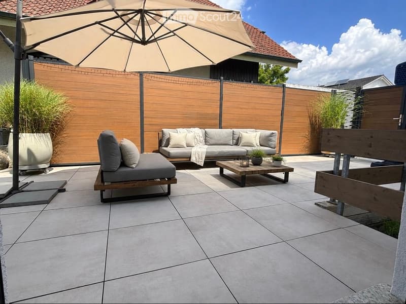 Outdoor patio with grey sofa, coffee table, lounge chair, umbrella, and small potted plants.