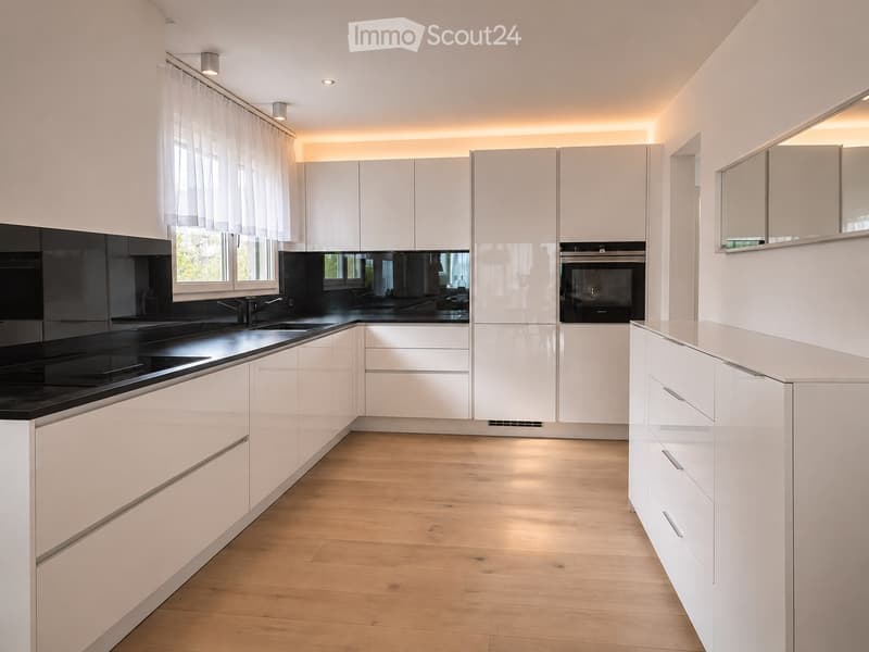 Modern kitchen with white cabinets, black countertops, and a built-in oven. Features include a large window with white curtains, sleek stainless steel fixtures, and a light wooden floor.