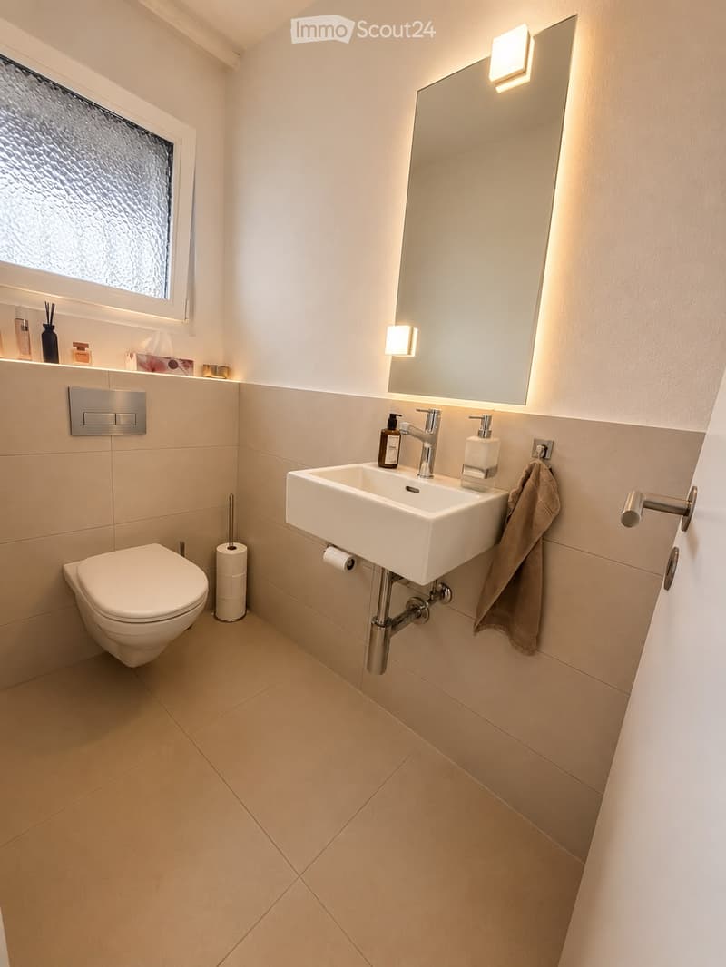 Modern bathroom with beige tiles, a white toilet, a white sink with a chrome faucet, a large mirror with built-in lighting, and a towel hanging on a chrome rack.