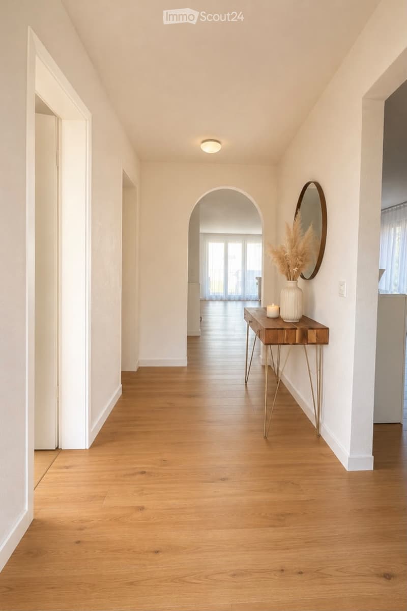 Bright hallway with wooden flooring, white walls, arched doorway leading to another room, small table with vase and candle, and a round mirror on the wall.