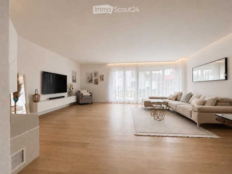 Modern living room with light wooden flooring, large windows with sheer curtains, a sectional sofa, coffee table, armchair, TV, and decorative items.