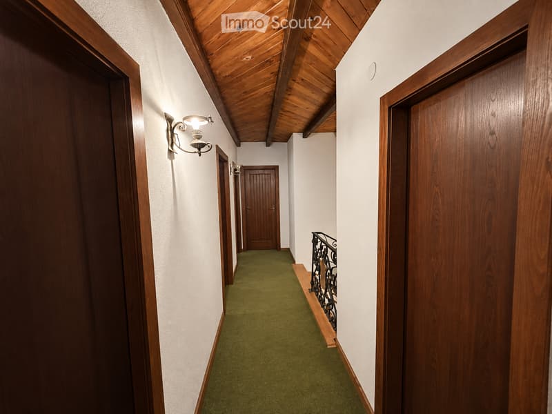 A narrow hallway with wooden doors, green carpeting, and a wooden ceiling. A wall-mounted light fixture is visible on the left side, and a staircase with a metal railing is seen on the right.