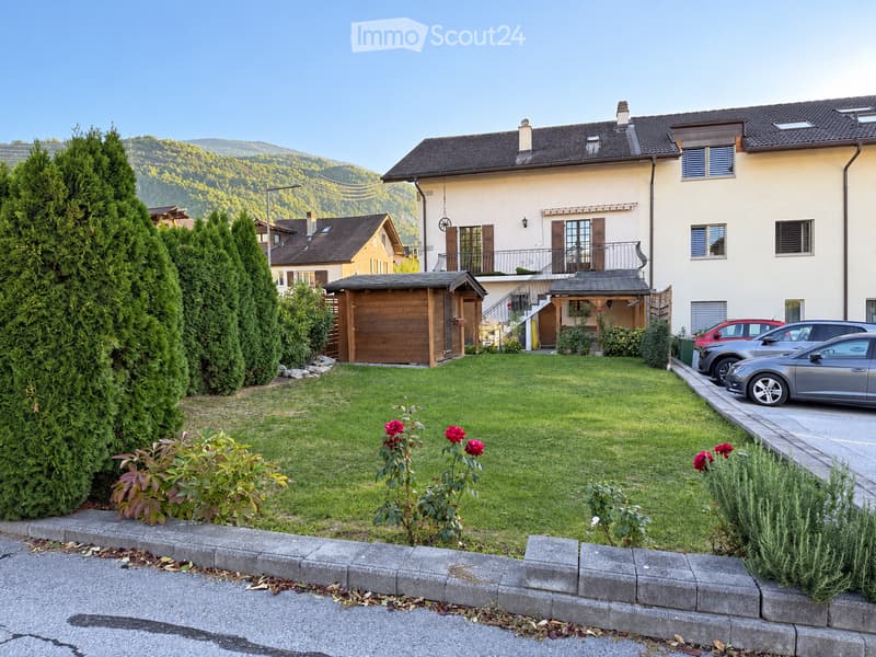 A well-maintained garden with a lawn, rose bushes, and tall evergreen trees, with a two-story house and a wooden garage in the background.