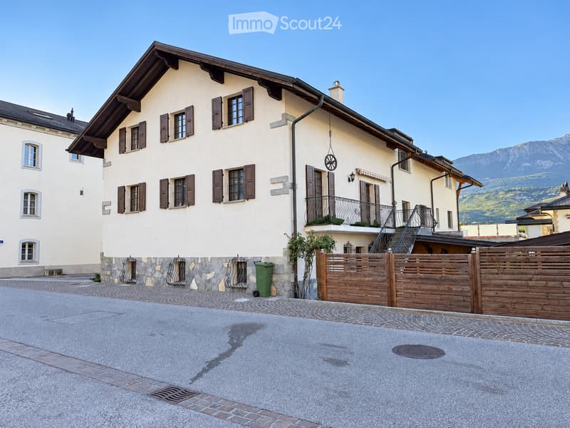 Two-story house with light yellow walls, dark brown shutters, and a balcony on the upper floor. The house features a stone base, a wooden fence, and a view of mountains in the background.