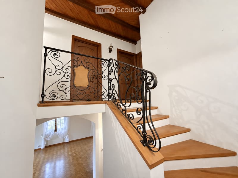 Staircase with wooden steps and ornate black wrought iron railing, leading to a room with wooden flooring and white walls.