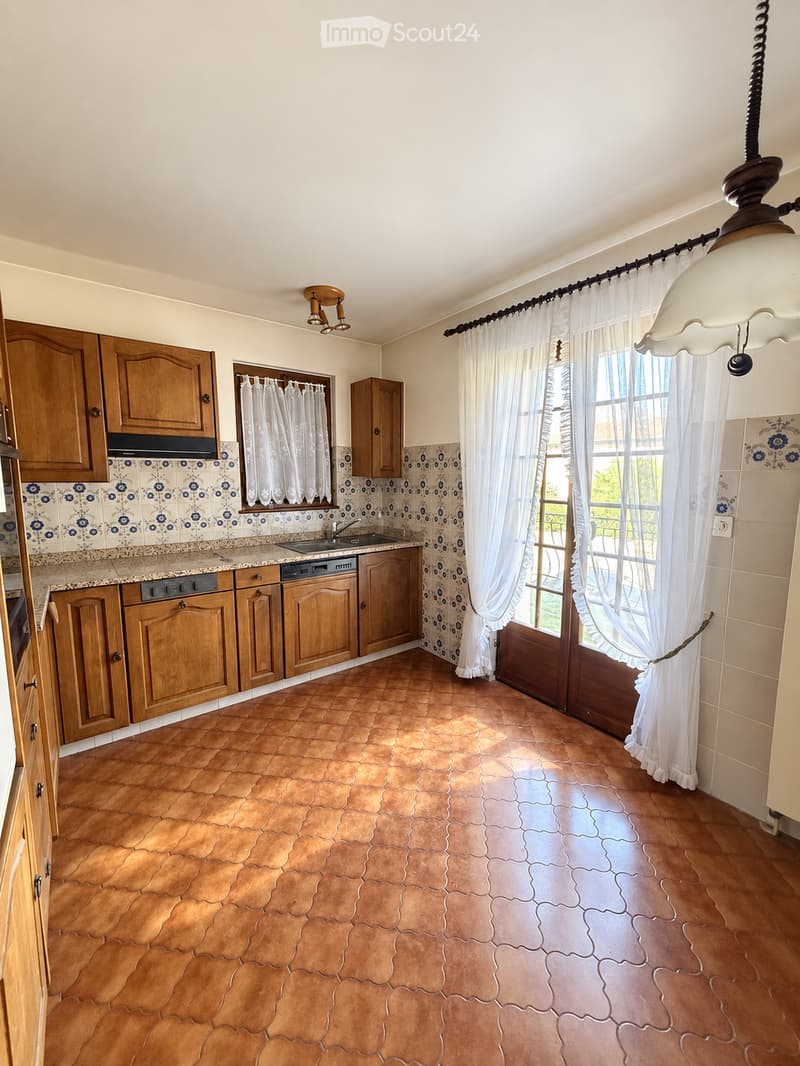 Bright kitchen with wooden cabinets, granite countertop, sink, dishwasher, and large windows with white curtains. The floor is made of terracotta tiles.
