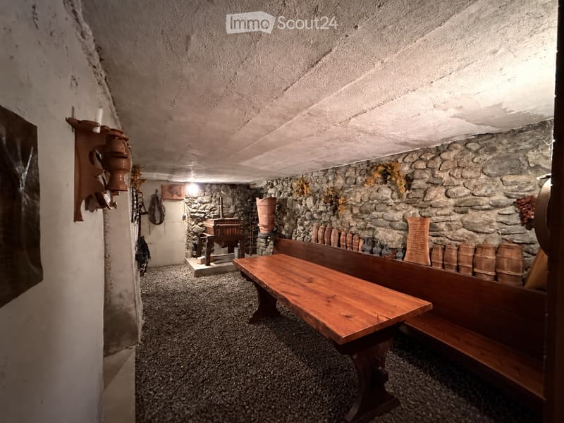 A rustic room with stone walls, wooden benches, a table, barrels, and a vase on the wall.