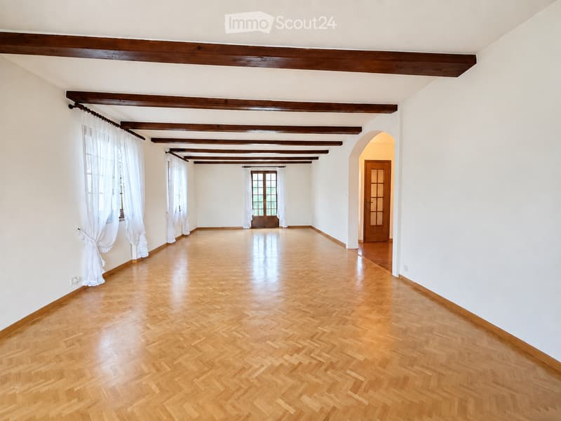 Empty room with wooden herringbone flooring, white walls, exposed wooden beams, and large windows with white curtains. No furniture or equipment present.