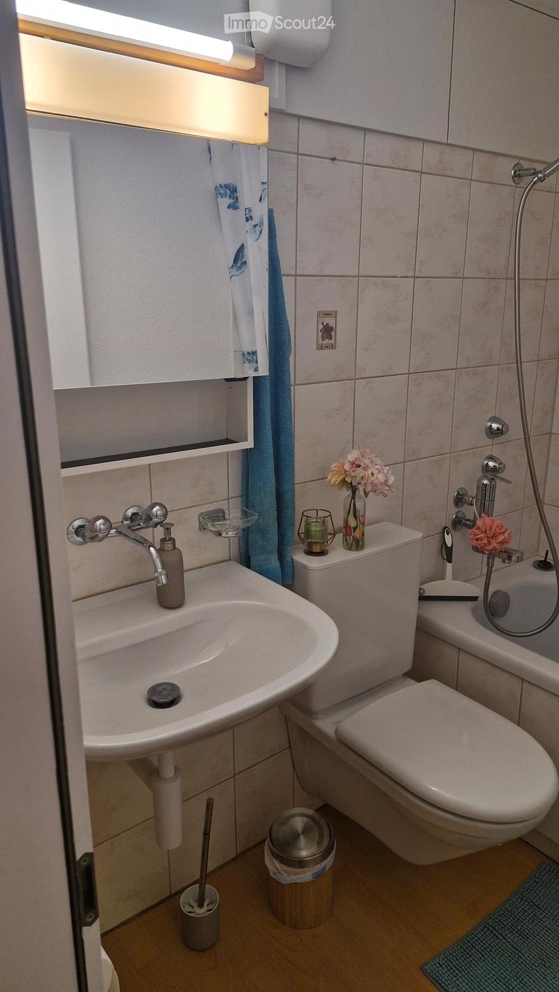 Modern bathroom with a white sink, toilet, bathtub, and showerhead. Features a mirror with a built-in shelf, a vase with flowers, and a candle on the toilet tank.