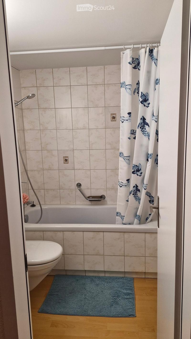 Bathroom with a bathtub, toilet, shower curtain, and a blue bath mat on the wooden floor.