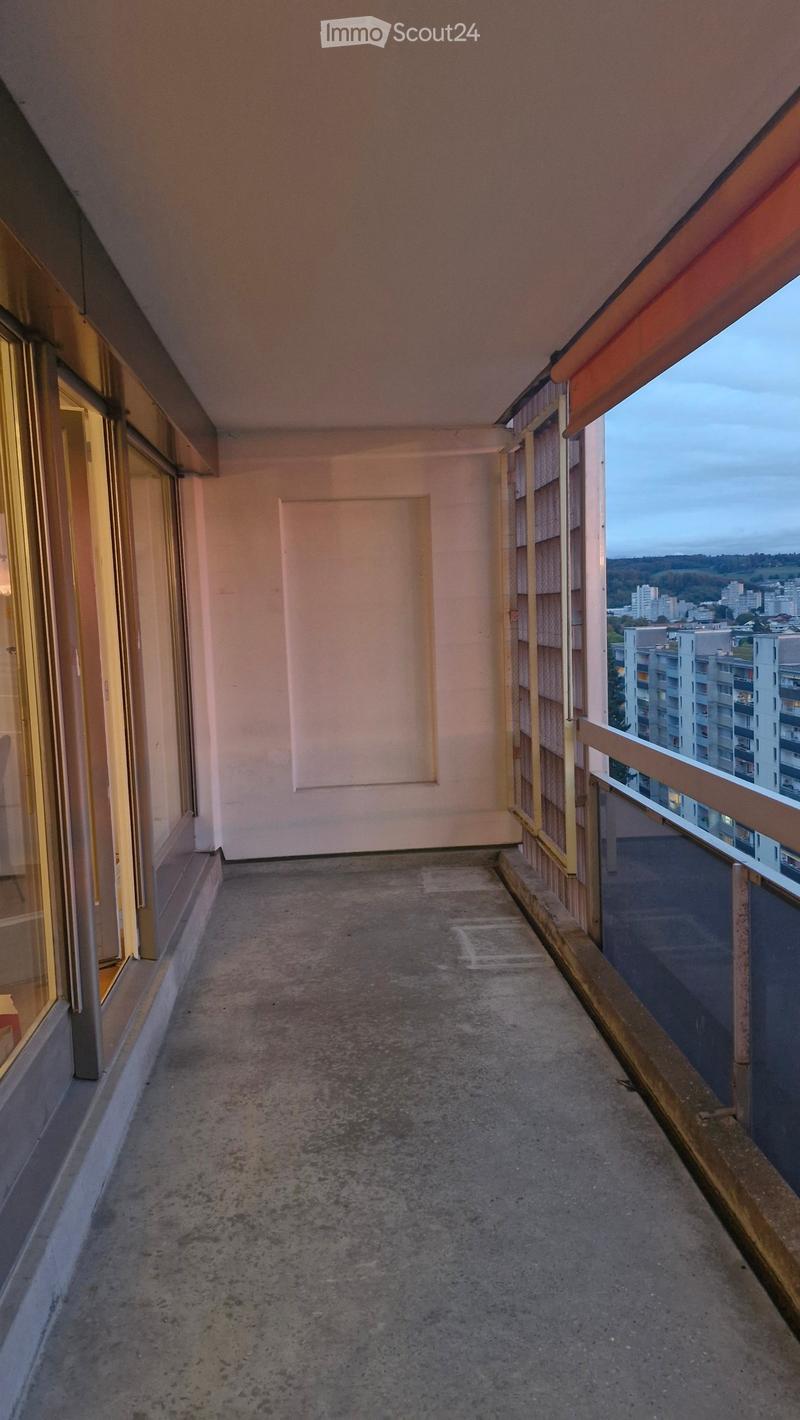 Empty balcony with concrete floor, metal railing, and large windows overlooking a cityscape.