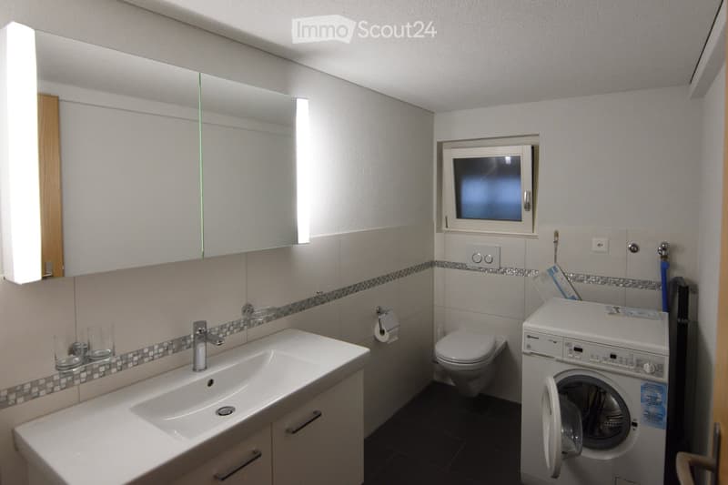 Modern bathroom with a white sink, mirror, toilet, and washing machine. The walls are tiled with a decorative border, and the floor is dark.