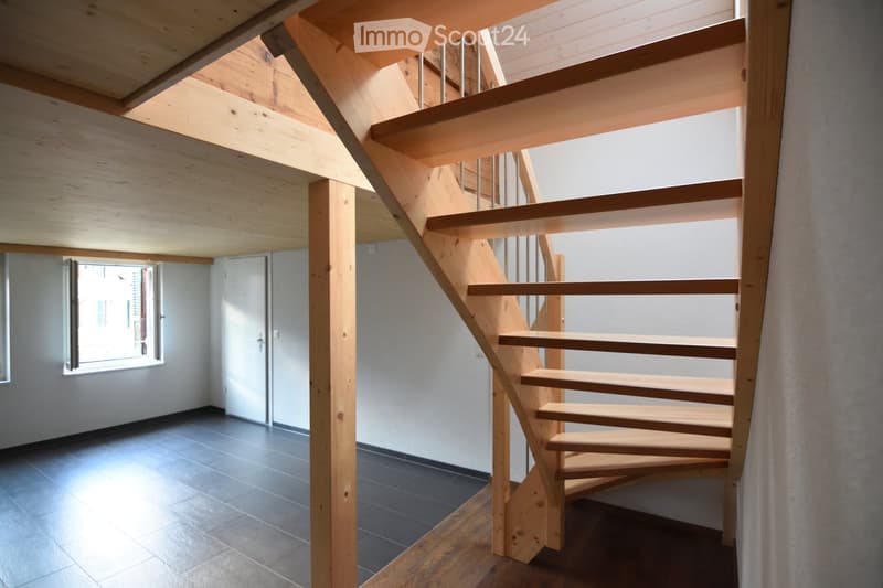 Modern wooden staircase with open wooden railings and metal balusters, leading to an upper level. The hallway has a tiled floor and white walls, with a window on the left side allowing natural light to enter.