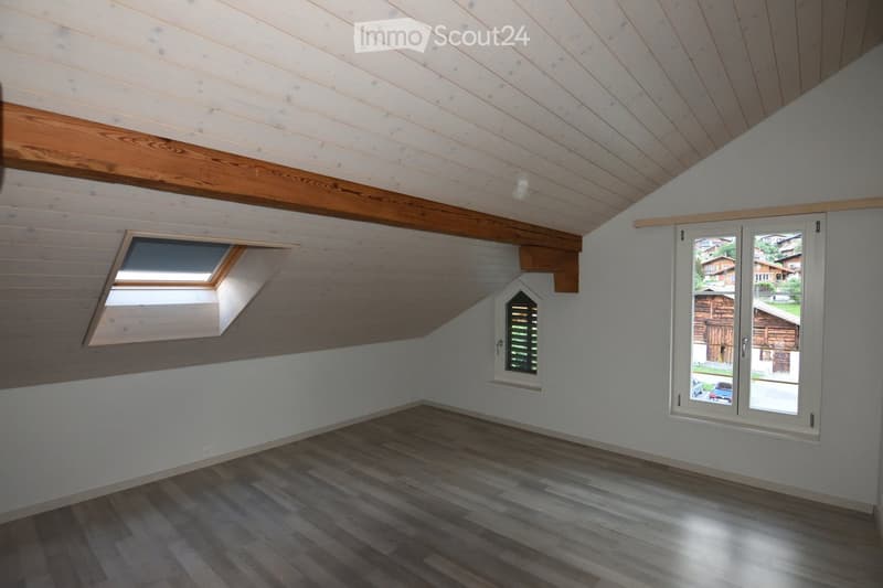 Empty attic room with wooden slat ceiling, exposed wooden beams, skylight window, and large window overlooking a wooden house exterior.