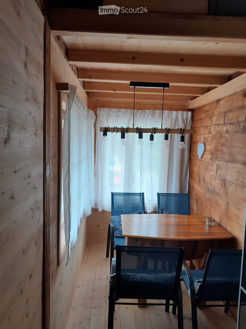 A small, cozy dining area with wooden walls and floor, featuring a wooden table and four black chairs, illuminated by a hanging light fixture with multiple bulbs.