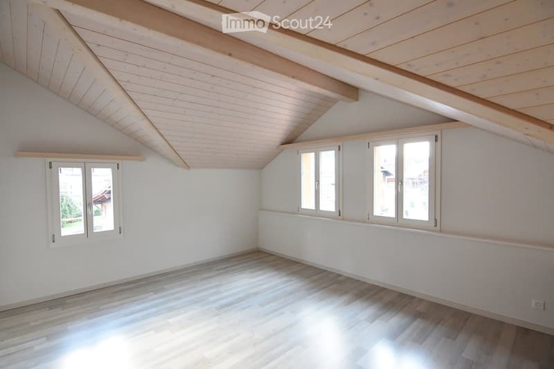 Empty room with wooden flooring, white walls, and exposed wooden beams on the ceiling. Two windows with white frames are present, one on each wall, allowing natural light to enter.