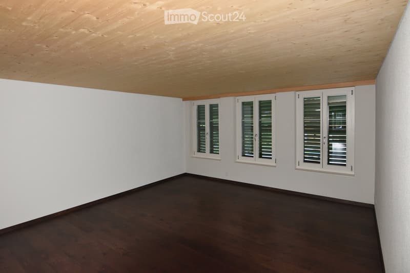 Empty room with white walls, dark wooden floor, and multiple windows with shutters. The ceiling is made of light wood panels.