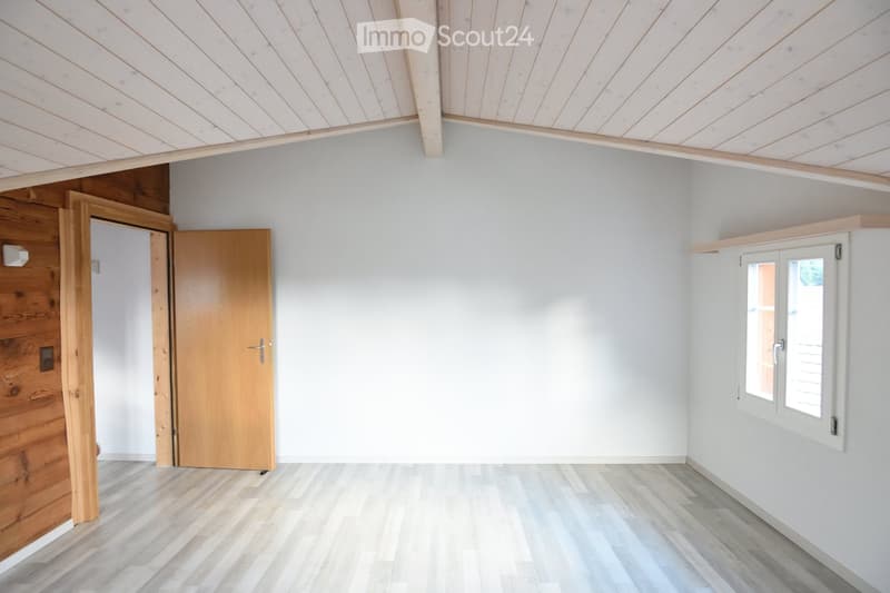 Empty room with light grey walls, wooden floor, and a window with white frame. There are two wooden doors on the left side, one leading to another room.