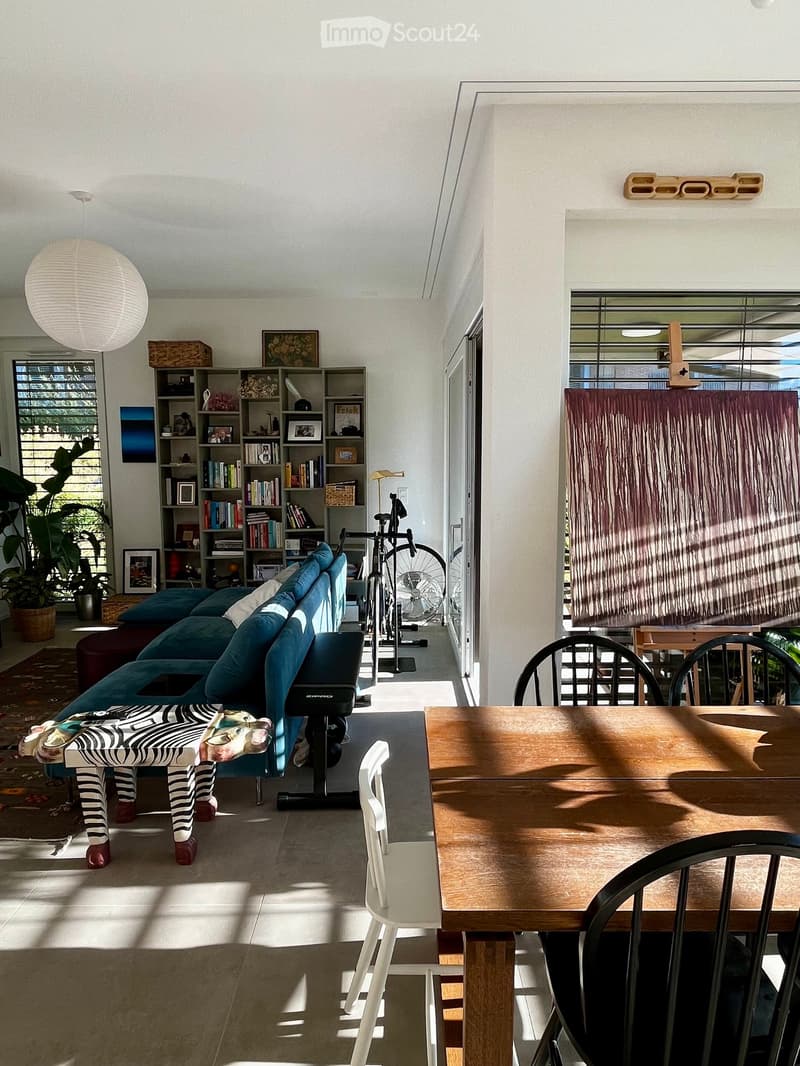 Modern living room with a blue sofa, wooden dining table, chairs, bookshelf, bicycle, and decorative items.