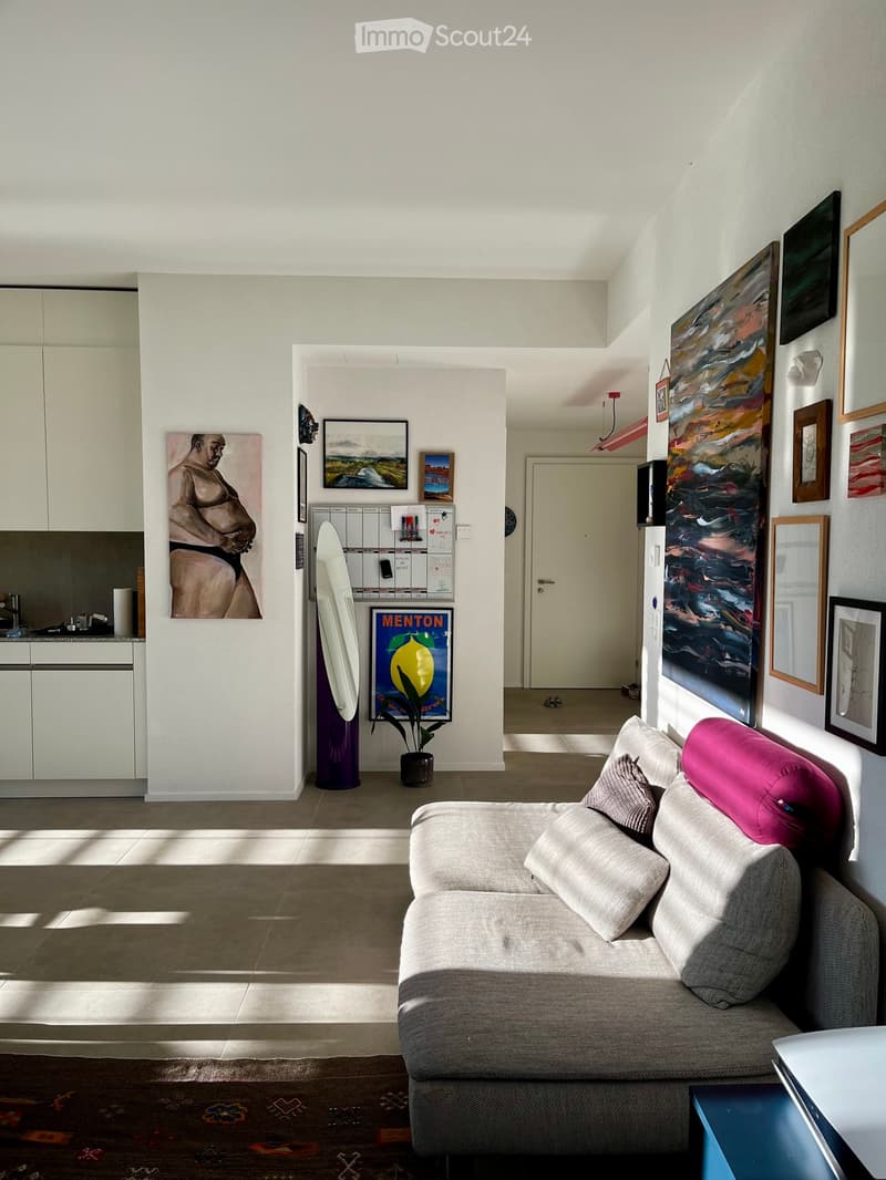 A modern living room with a grey sofa, a small table, white cabinets, and several framed artworks on the walls. A potted plant sits near the doorway.