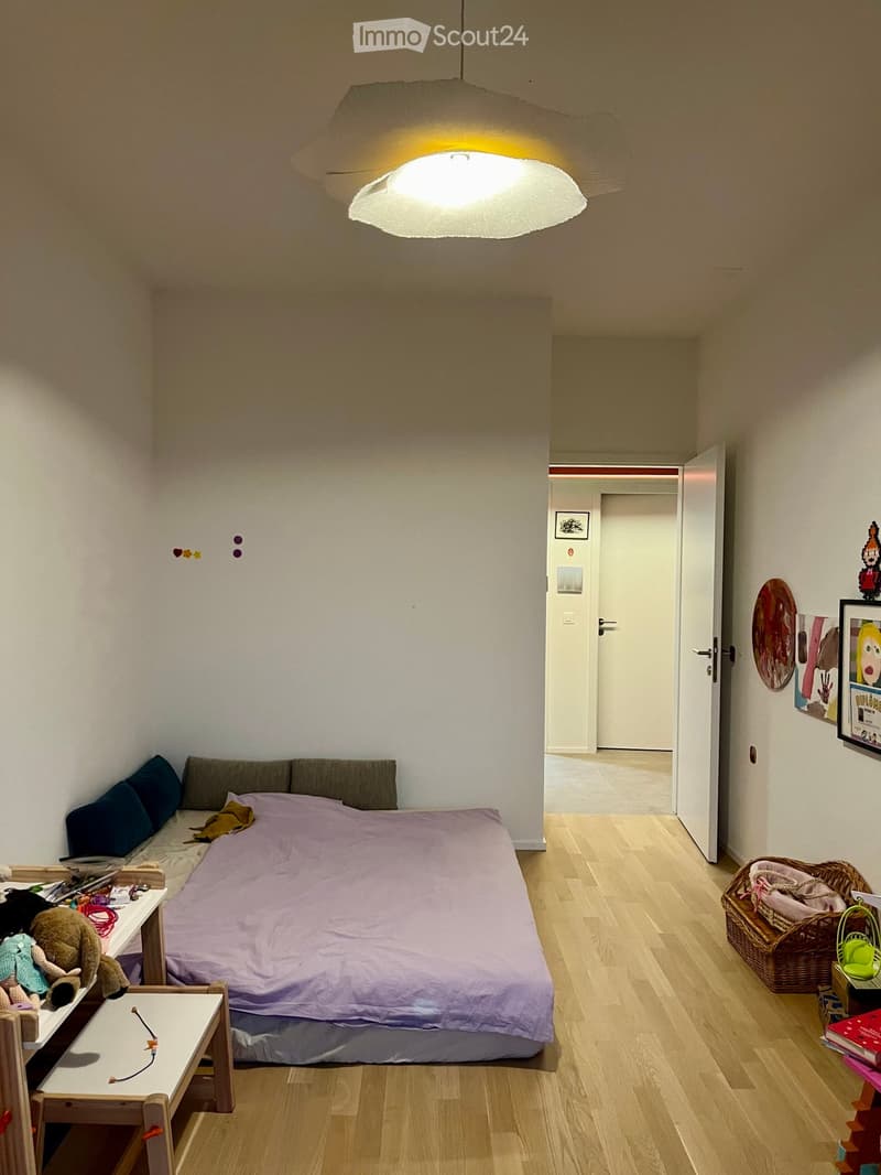 A small, cozy bedroom with a single bed, a small sofa, a wooden table with toys, a wicker basket, and a colorful wall decoration.