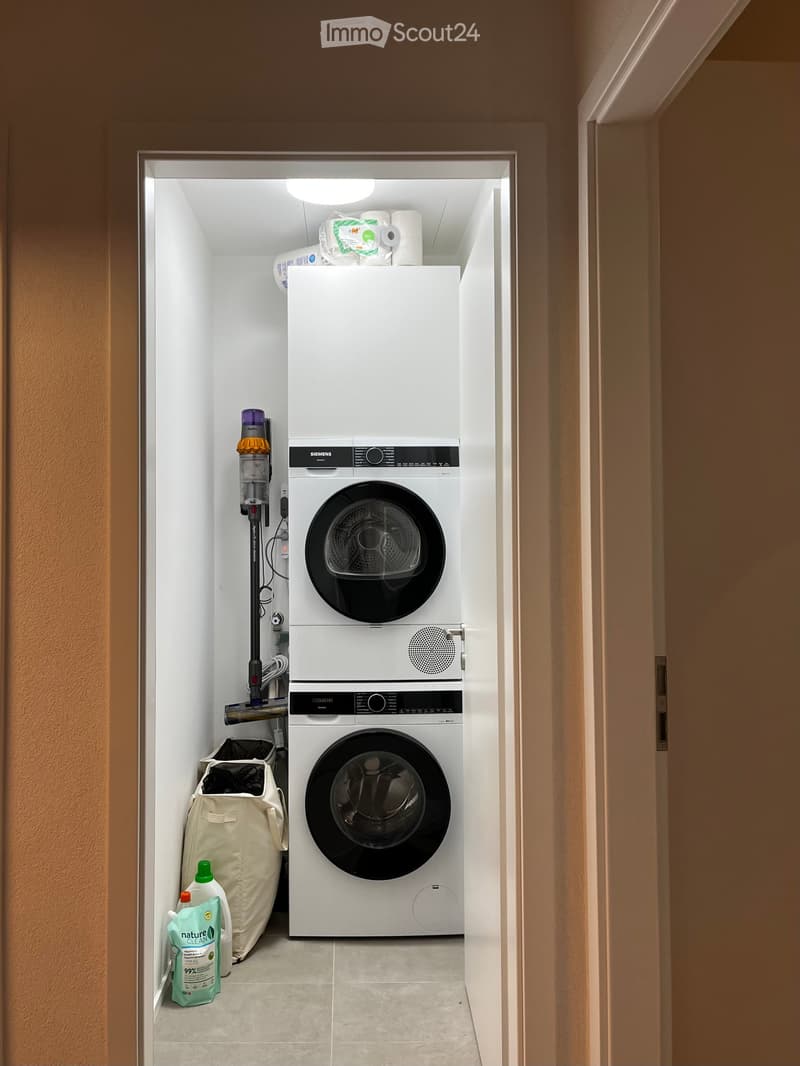Compact bathroom with a stacked washing machine and dishwasher unit, vacuum cleaner, laundry bag, and cleaning supplies.
