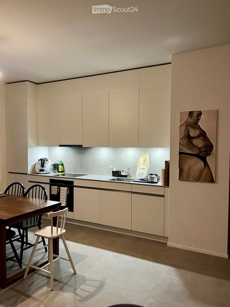Modern kitchen with light-colored cabinets, a wooden dining table with chairs, and various appliances including a blender and oven. A painting is displayed on the wall.
