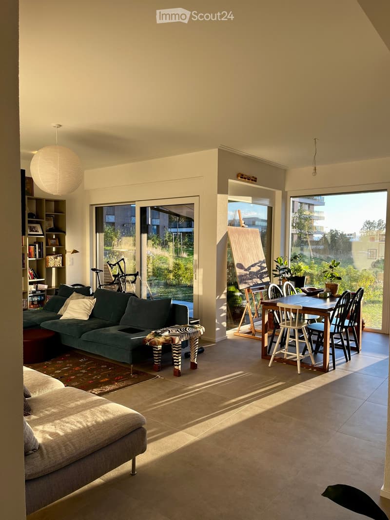 Bright living room with large windows and a view of greenery, featuring a green sectional sofa, a wooden dining table with chairs, a shelf with books and decor, and a bicycle in the corner.