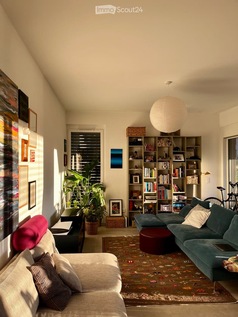 Living room with two sofas, a round red stool, a bookshelf, a coffee table, and a large plant near the window. The room features a colorful rug and modern decor.