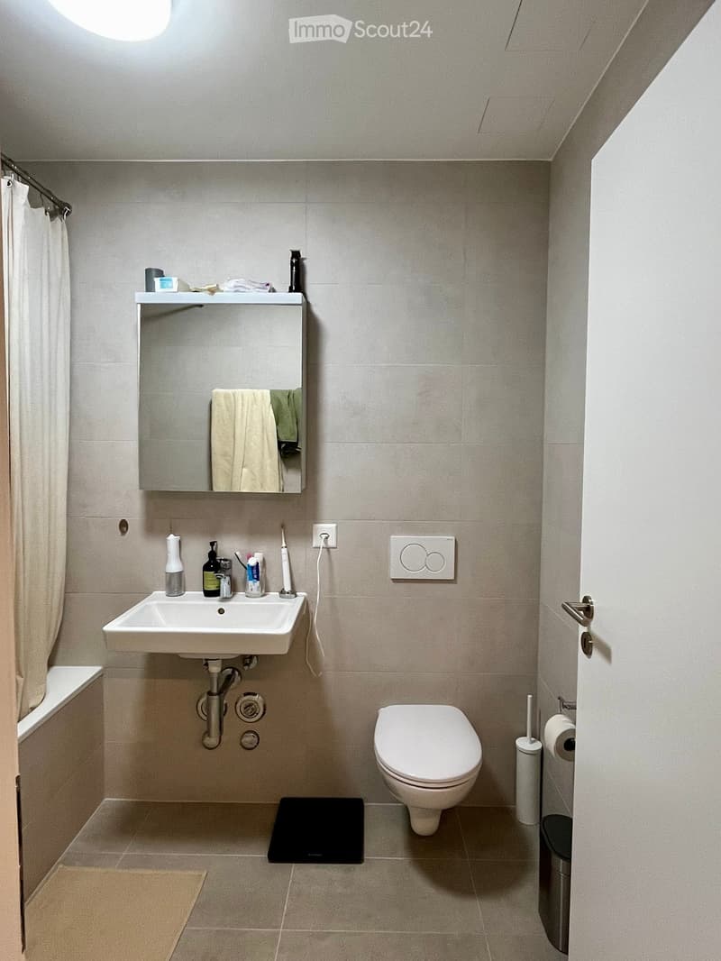 Modern bathroom with a white sink, toilet, mirror, and various toiletries. Features a beige tiled floor and walls, a shower curtain, and a small black mat in front of the toilet.