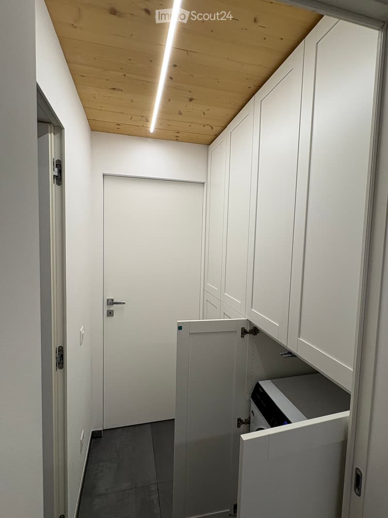 A narrow hallway with white walls, a wooden ceiling, and built-in white cabinets. A door is visible at the end, and a private washing machine is partially visible in an open cabinet.