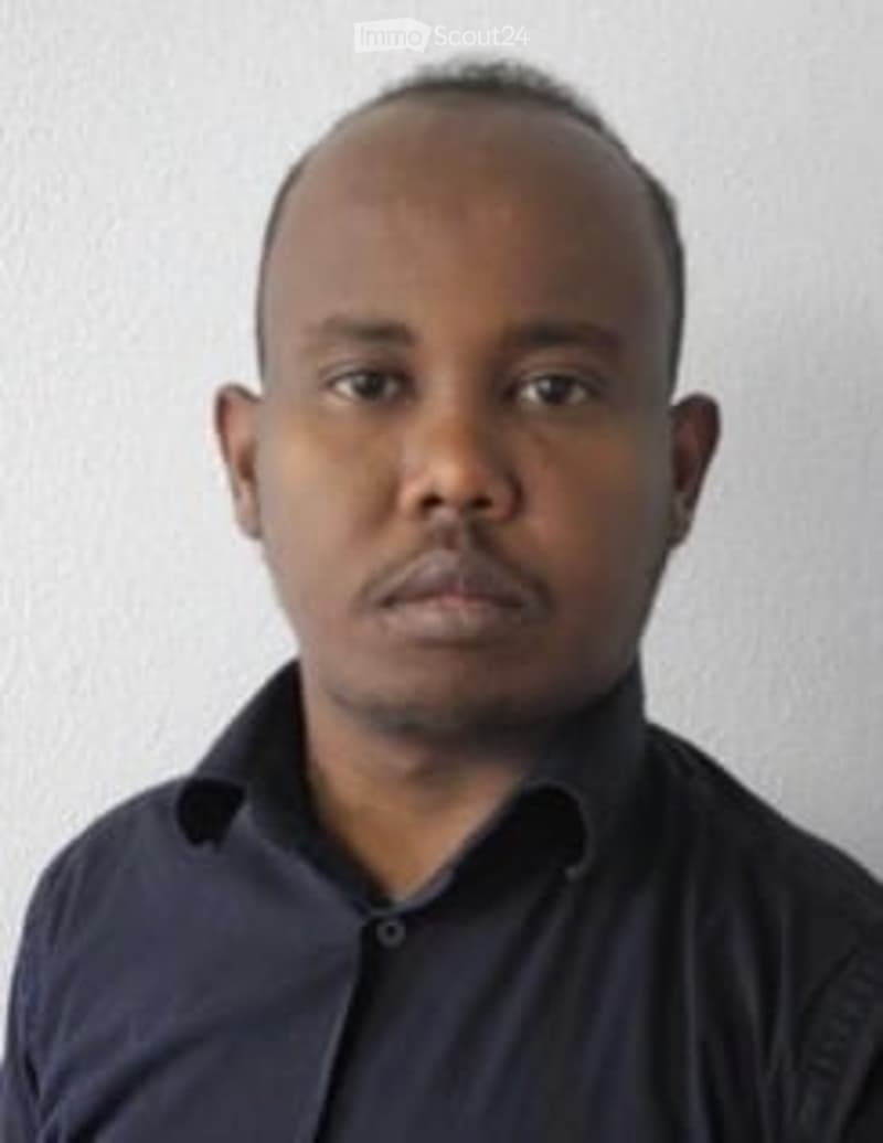 A man in a dark blue shirt stands against a plain white background. He has short hair and a neutral expression.
