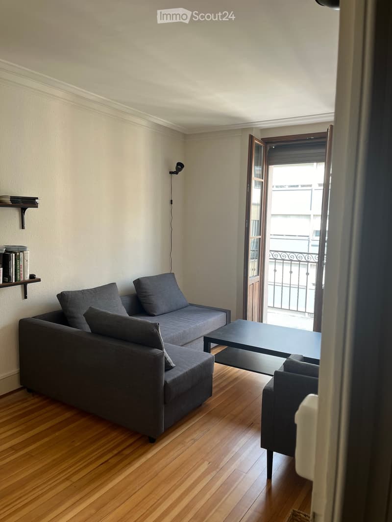 Living room with wooden flooring, grey sofa, small table, and a shelf with books.