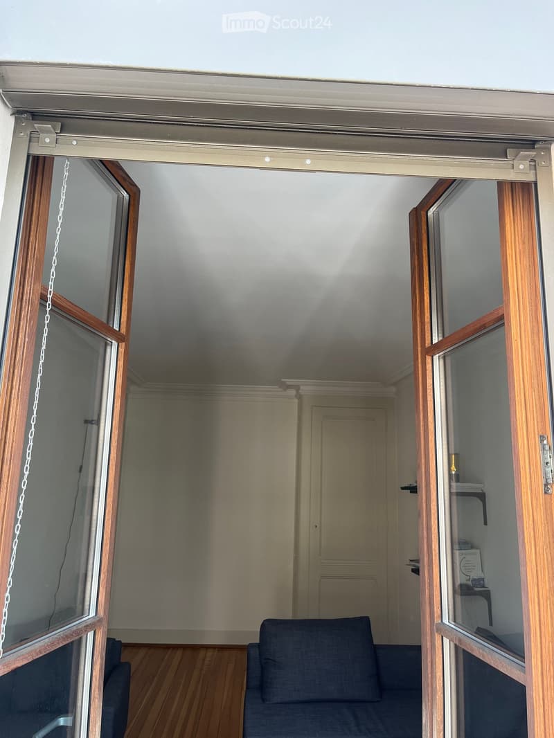 A small, unfurnished room with wooden flooring, a single sofa, and large sliding glass doors leading to an outdoor area.
