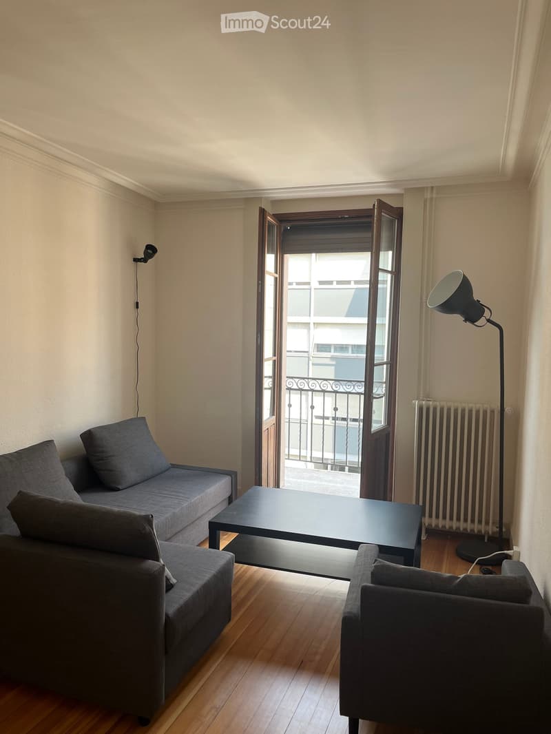 A modern living room with a grey sofa, a black coffee table, a single armchair, and a floor lamp. The room features wooden flooring and an open door leading to a balcony.