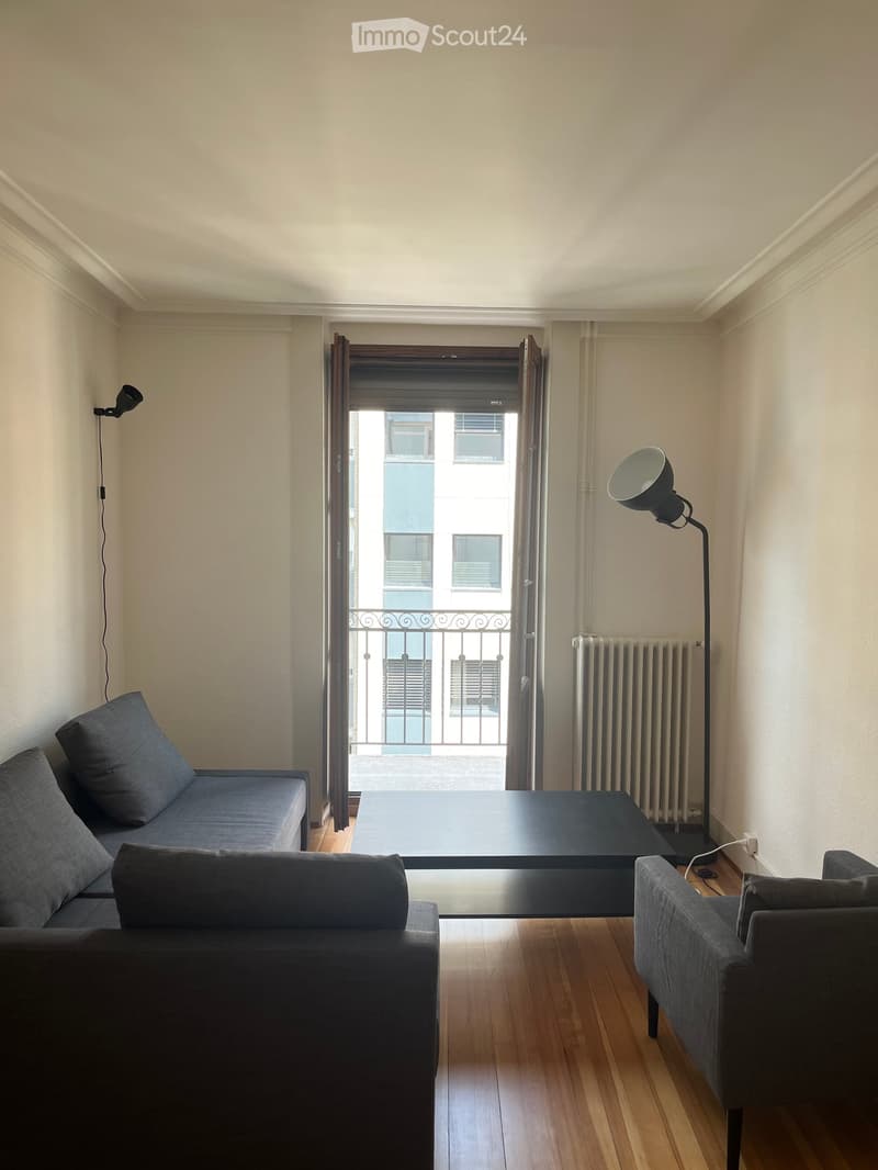 Modern living room with a sofa, coffee table, armchair, floor lamp, radiator, and an open balcony door leading to a view of the city.