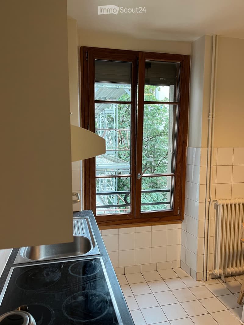 Empty kitchen with tiled walls and floor, electric stove, sink, and radiator. Large window offers a view of a balcony and greenery.