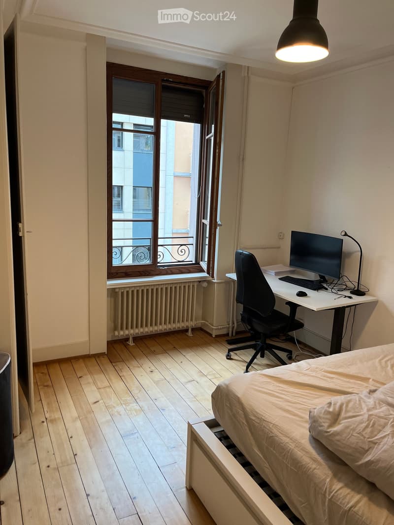 Cozy bedroom with wooden flooring, a bed with white bedding, a desk with a computer, chair, and lamp, and a window with a view of the city.