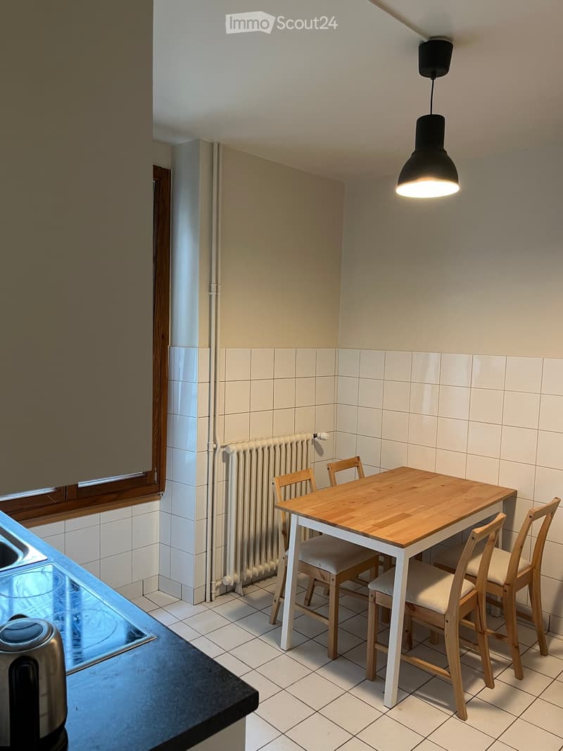 A small kitchen with a wooden dining table and four chairs, a radiator, and a hanging lamp. The walls are tiled, and a countertop is visible on the left side.