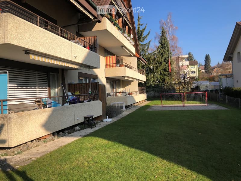 Well-maintained garden with green lawn, small seating area, and soccer goal. The building features balconies, awnings, and outdoor furniture.