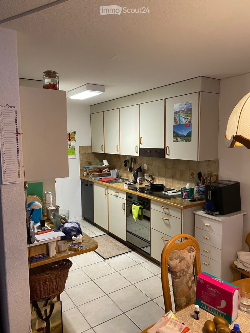 The kitchen features white cabinets, a tiled floor, and various appliances including a refrigerator, oven, dishwasher, and coffee maker. There is a wooden chair, a small table with items on it, and a wicker basket on the floor.
