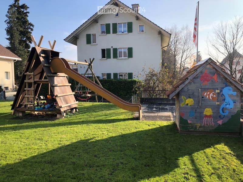A well-maintained garden with a wooden play structure featuring a slide and climbing elements, a small playhouse with ocean-themed artwork, and a two-story house with green shutters in the background.