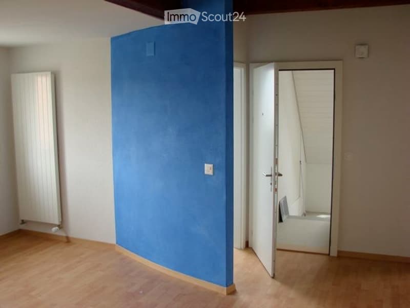 Empty room with wooden flooring, white walls, a blue partition, and an open doorway leading to a small bathroom.