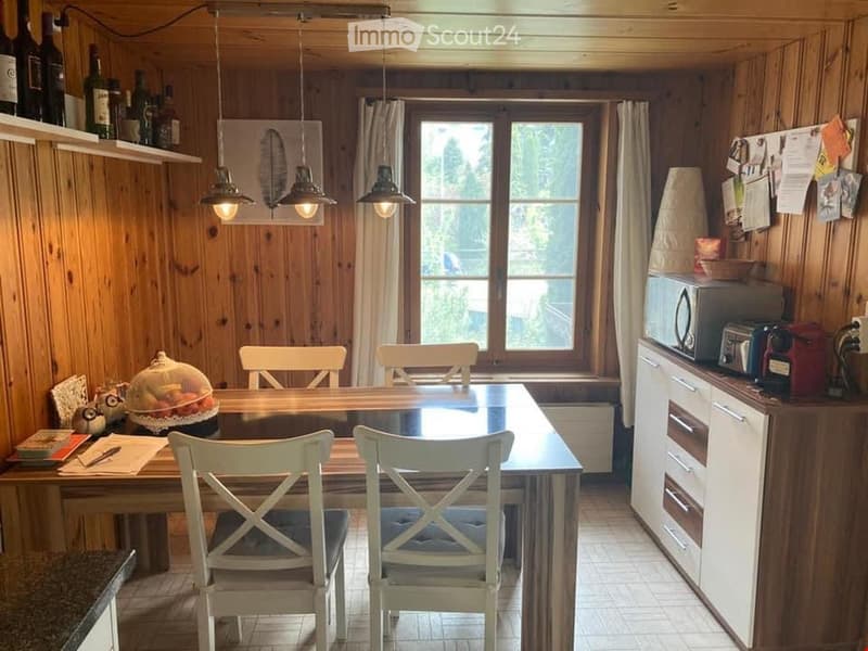 Cozy wooden-paneled room with a dining table and chairs, a small kitchen area with a microwave, toaster, and cupboard, and shelves holding wine bottles and a framed picture. Natural light streams in through large windows with curtains.