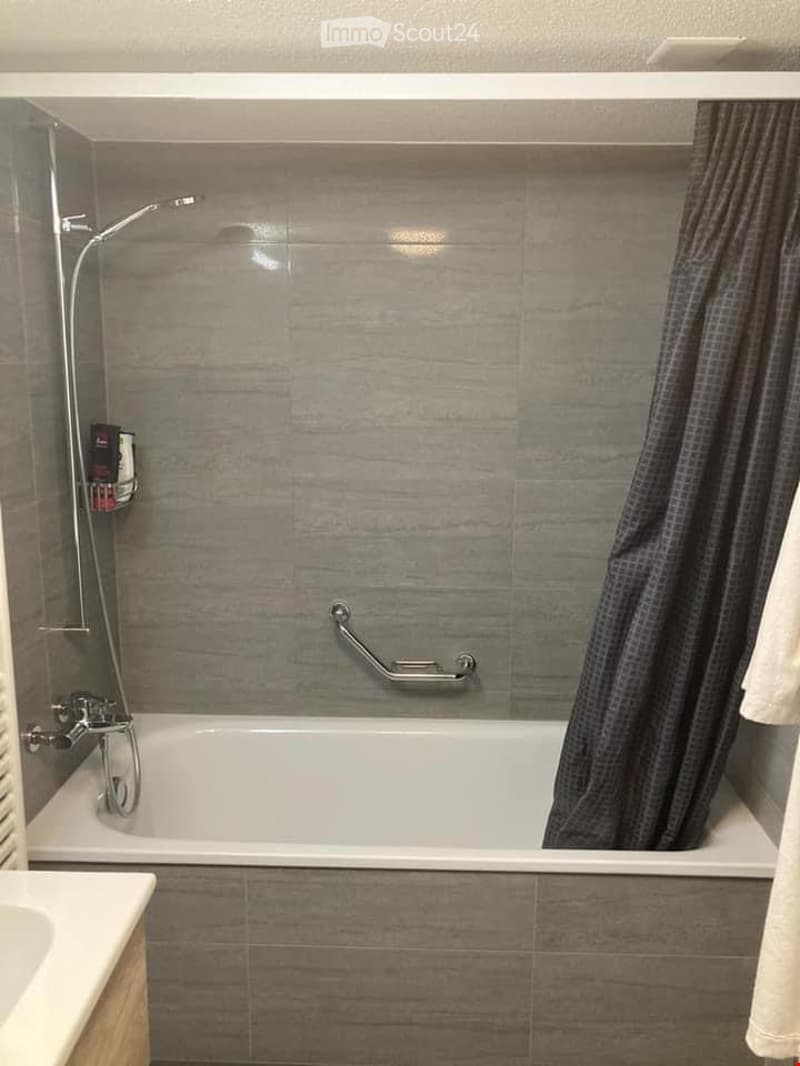 Modern bathroom with a bathtub, showerhead, and a handrail for safety. The walls are tiled in a grey marble pattern, and a shower curtain is partially visible.