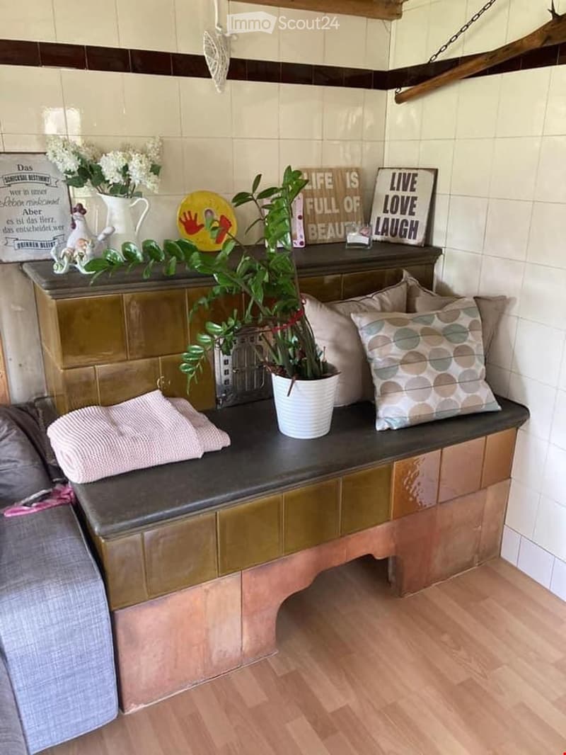 A cozy, furnished room with a wooden bench, potted plant, decorative pillows, and a vase with flowers on top. The bench has a dark surface and is adorned with various decorative items, including signs and a framed picture.
